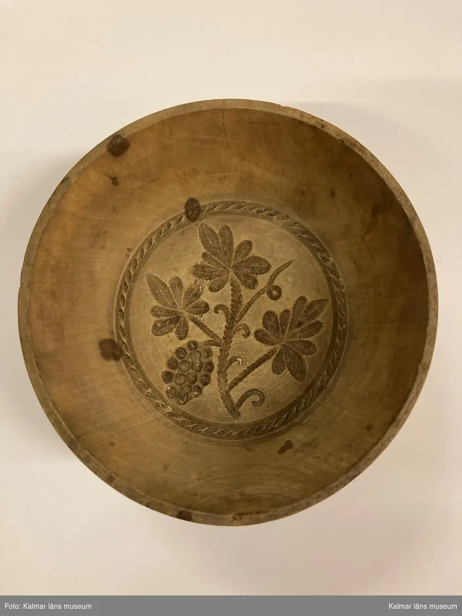 KLM 6153. Ostform. Av trä. Svarvad skål med låg fot. I botten ett snidat mönster i form av en vinkvist med en druvklase.