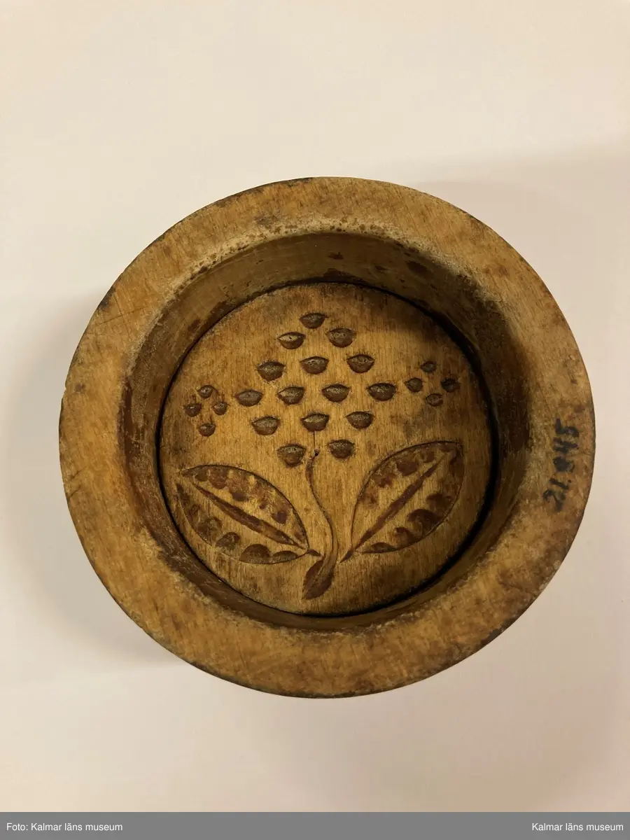 KLM 21845. Smörform. Av trä, bestående av två delar. Den ena är en platta med skuret blomornament, på denna har det baktill ursprungligen suttit ett handtag. Den andra delen är i form av en cylindrisk hylsa.