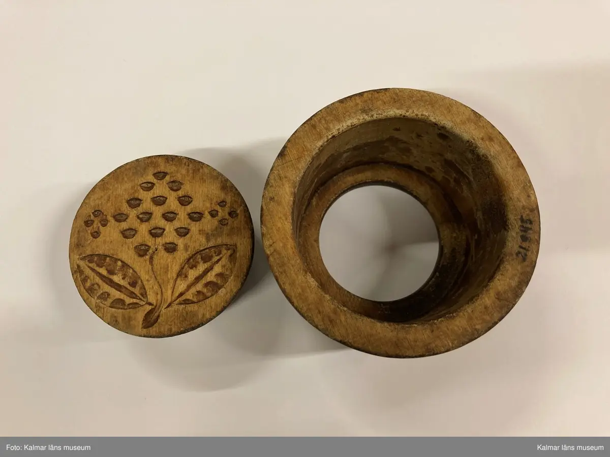 KLM 21845. Smörform. Av trä, bestående av två delar. Den ena är en platta med skuret blomornament, på denna har det baktill ursprungligen suttit ett handtag. Den andra delen är i form av en cylindrisk hylsa.