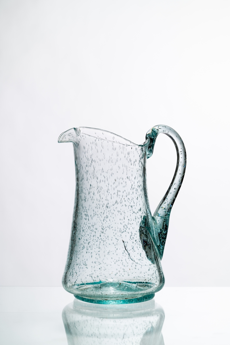 Kanna, den så kallade Kungakannan, av ljust grönfärgat glas. Kannan har rund botten med puntelmärke, insvängd form upp mot mynningen, försedd med grepe och utvikt snip. Spricka i glaset nedanför grepen. Bubblorna i glaset beror på att glasmassan inte kom upp i tillräckligt hög temperatur, för att blåsorna i den tröga massan helt skulle kunna försvinna. Hantverksmässigt tillverkad. Se vidare Historik.