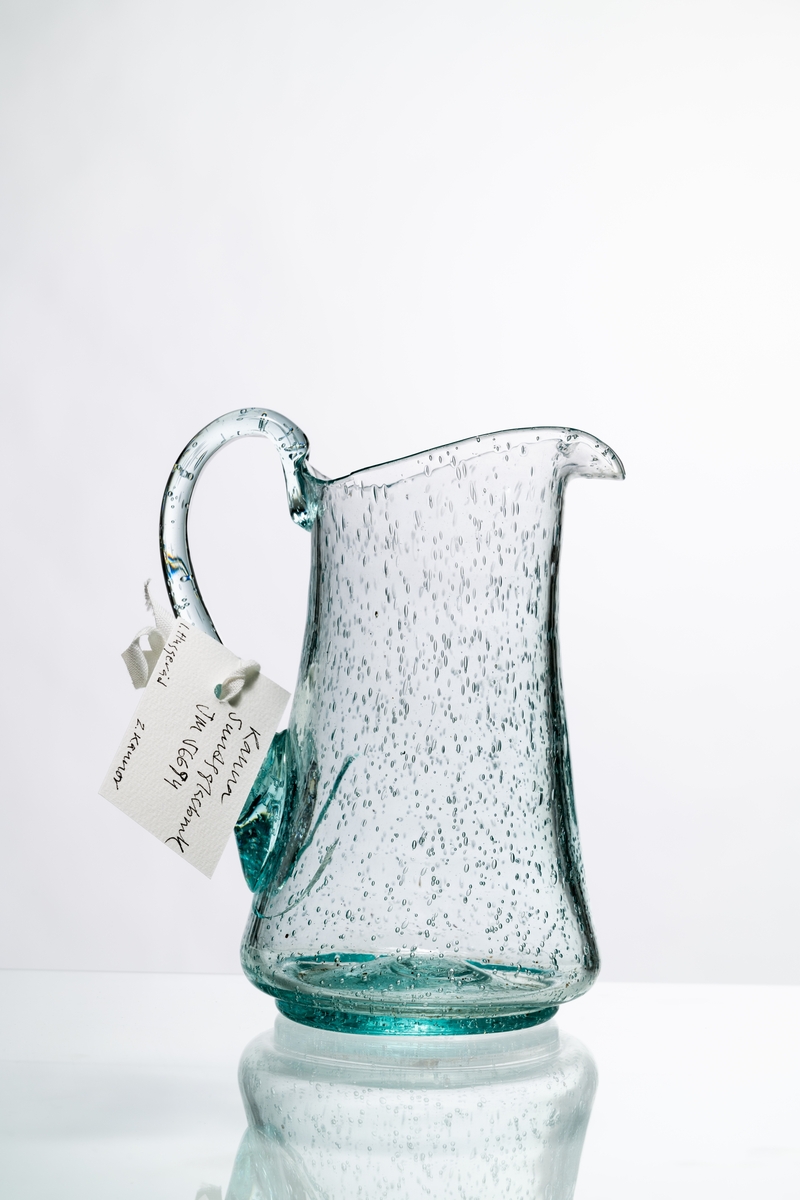 Kanna, den så kallade Kungakannan, av ljust grönfärgat glas. Kannan har rund botten med puntelmärke, insvängd form upp mot mynningen, försedd med grepe och utvikt snip. Spricka i glaset nedanför grepen. Bubblorna i glaset beror på att glasmassan inte kom upp i tillräckligt hög temperatur, för att blåsorna i den tröga massan helt skulle kunna försvinna. Hantverksmässigt tillverkad. Se vidare Historik.