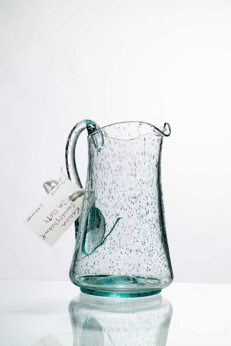 Kanna, den så kallade Kungakannan, av ljust grönfärgat glas. Kannan har rund botten med puntelmärke, insvängd form upp mot mynningen, försedd med grepe och utvikt snip. Spricka i glaset nedanför grepen. Bubblorna i glaset beror på att glasmassan inte kom upp i tillräckligt hög temperatur, för att blåsorna i den tröga massan helt skulle kunna försvinna. Hantverksmässigt tillverkad. Se vidare Historik.