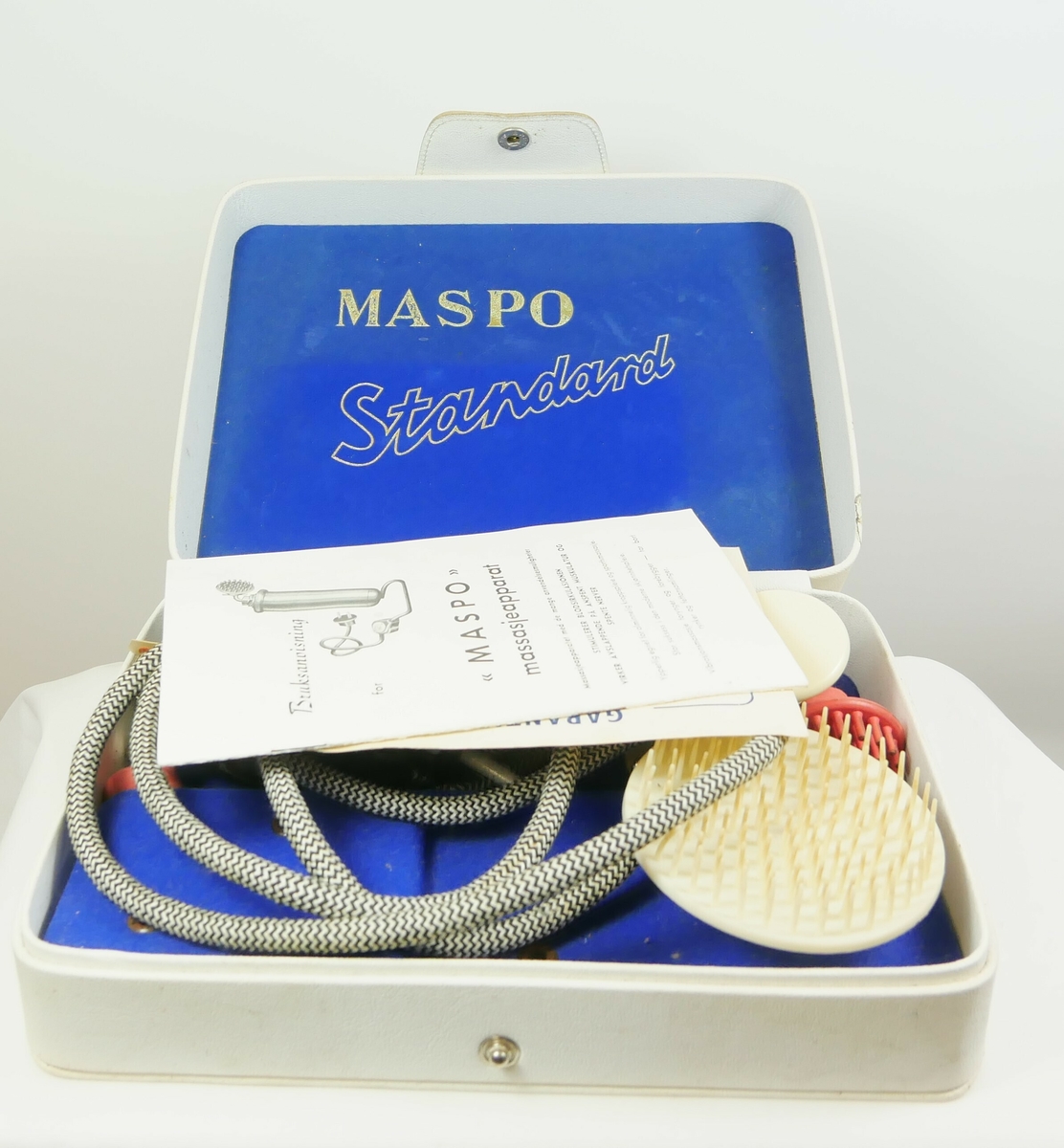 Hvit rektangulær boks med lokk. Inneholder et massasjeapparat. 