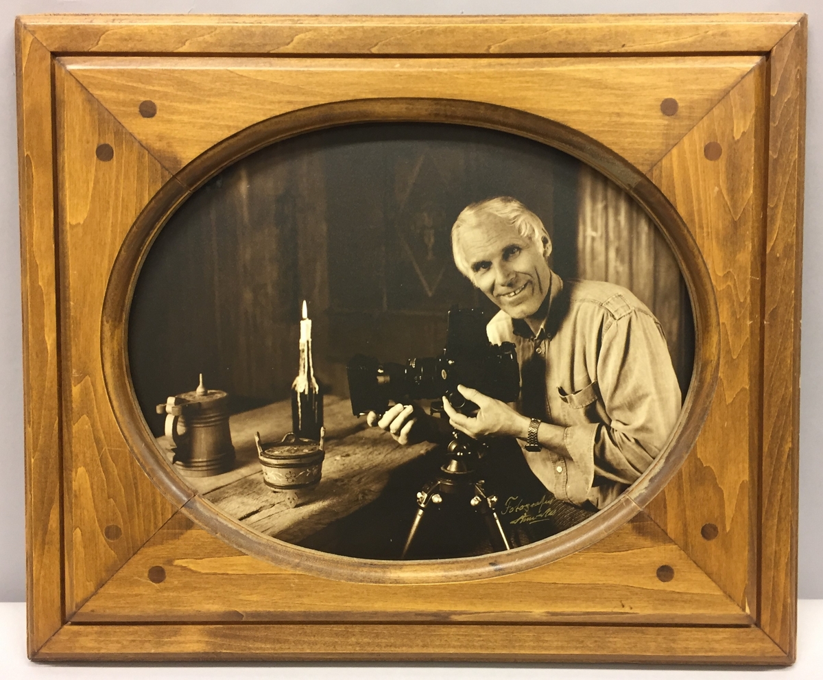 Bilete i treramme. Portrett av fotografen Arne Aas.Tekst bak på biletet:"Arne i den gamle stoga i studioet sitt i 1987".