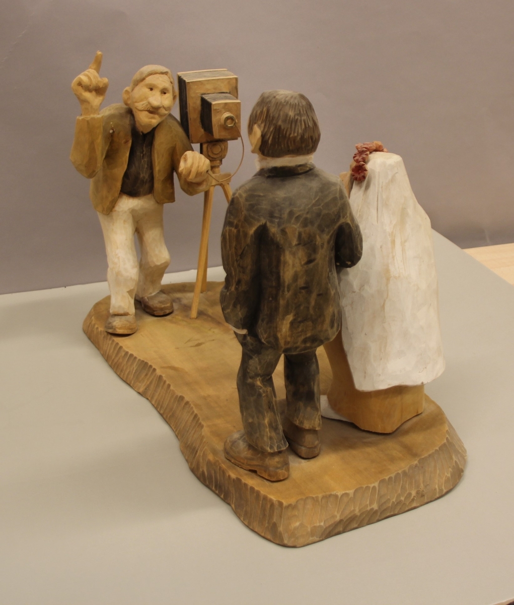 Skulptur som syner Arne Aas som tek bilete av eit brudepar. Skulpturen er skoren i tre.Tekst under:"Bjarne Walle fra Bamle laget denne gruppa på bestilling i fra fotografen Arne Aas i 1984" "Motiv: Arne Aas fotograferer brudepar". Bokstavane BW skore inn under skulpturen.