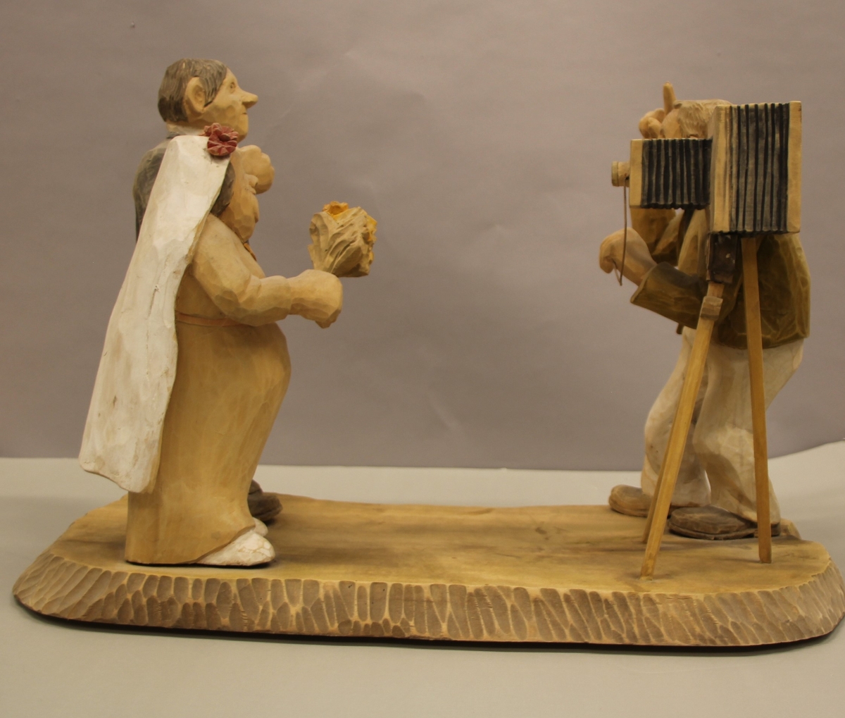 Skulptur som syner Arne Aas som tek bilete av eit brudepar. Skulpturen er skoren i tre.Tekst under:"Bjarne Walle fra Bamle laget denne gruppa på bestilling i fra fotografen Arne Aas i 1984" "Motiv: Arne Aas fotograferer brudepar". Bokstavane BW skore inn under skulpturen.