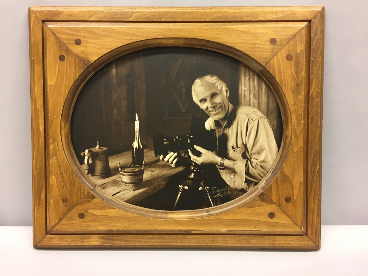 Bilete i treramme. Portrett av fotografen Arne Aas.Tekst bak på biletet:"Arne i den gamle stoga i studioet sitt i 1987".