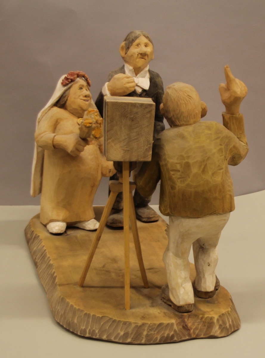 Skulptur som syner Arne Aas som tek bilete av eit brudepar. Skulpturen er skoren i tre.Tekst under:"Bjarne Walle fra Bamle laget denne gruppa på bestilling i fra fotografen Arne Aas i 1984" "Motiv: Arne Aas fotograferer brudepar". Bokstavane BW skore inn under skulpturen.