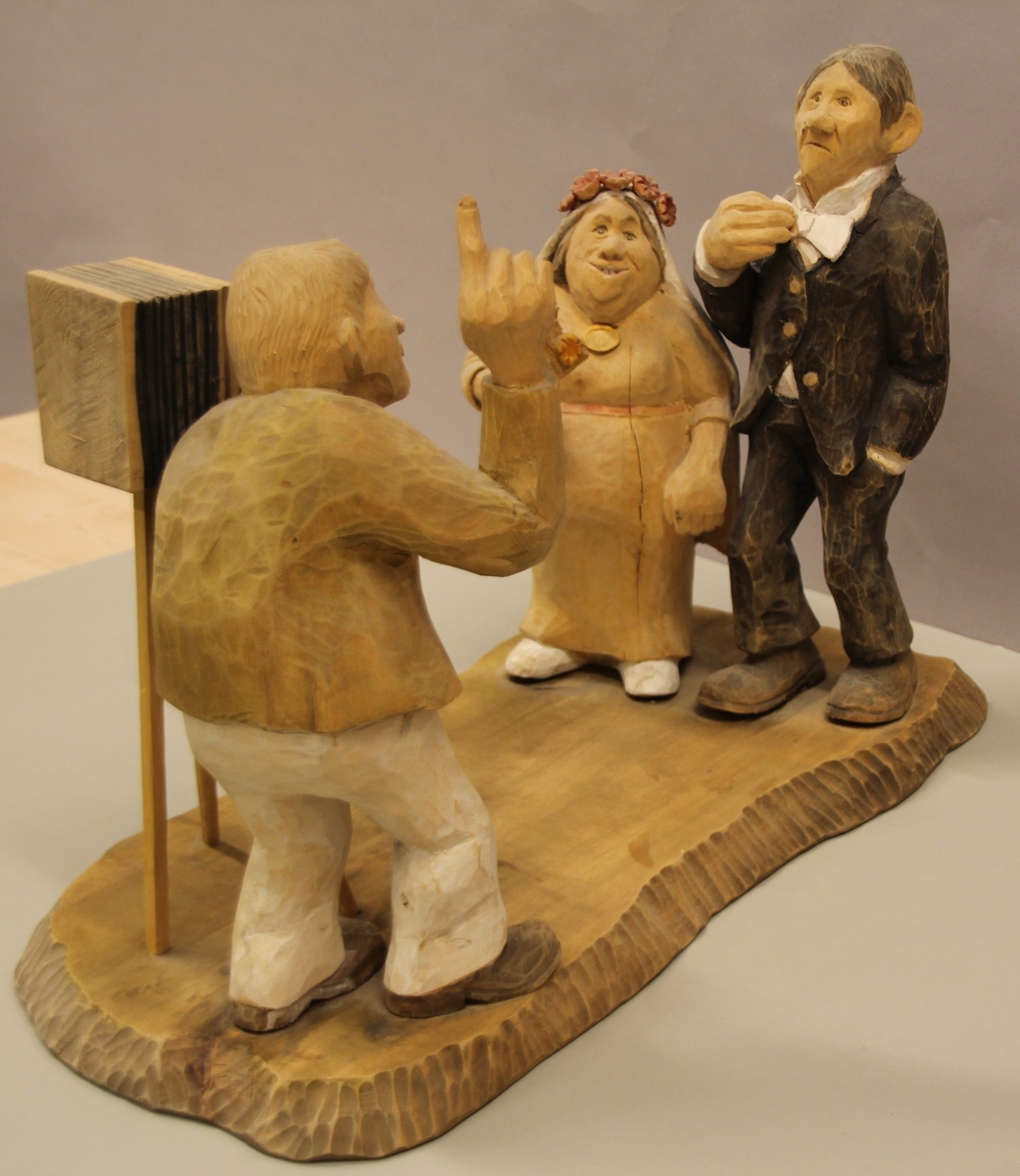 Skulptur som syner Arne Aas som tek bilete av eit brudepar. Skulpturen er skoren i tre.Tekst under:"Bjarne Walle fra Bamle laget denne gruppa på bestilling i fra fotografen Arne Aas i 1984" "Motiv: Arne Aas fotograferer brudepar". Bokstavane BW skore inn under skulpturen.