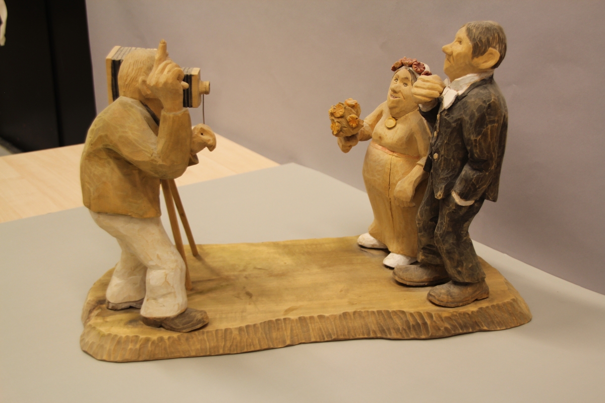 Skulptur som syner Arne Aas som tek bilete av eit brudepar. Skulpturen er skoren i tre.Tekst under:"Bjarne Walle fra Bamle laget denne gruppa på bestilling i fra fotografen Arne Aas i 1984" "Motiv: Arne Aas fotograferer brudepar". Bokstavane BW skore inn under skulpturen.
