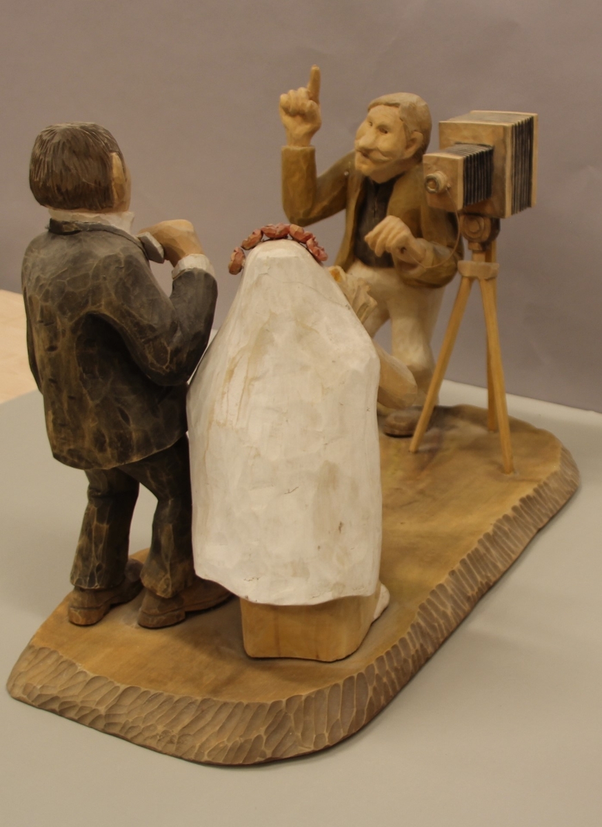 Skulptur som syner Arne Aas som tek bilete av eit brudepar. Skulpturen er skoren i tre.Tekst under:"Bjarne Walle fra Bamle laget denne gruppa på bestilling i fra fotografen Arne Aas i 1984" "Motiv: Arne Aas fotograferer brudepar". Bokstavane BW skore inn under skulpturen.