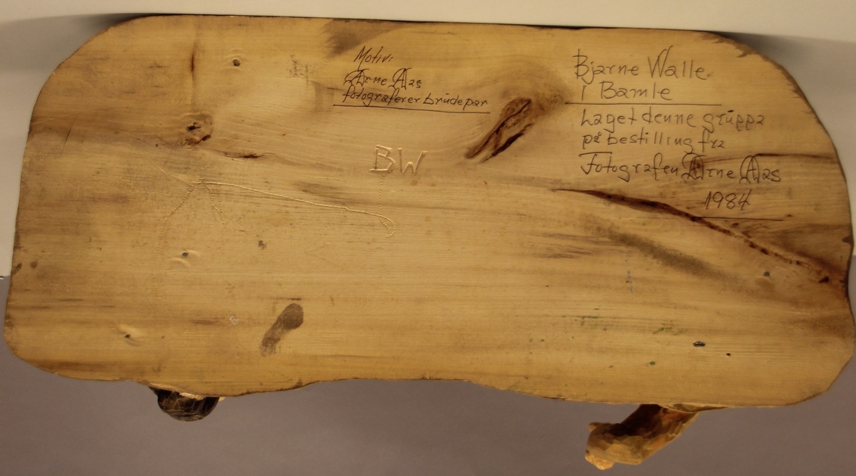 Skulptur som syner Arne Aas som tek bilete av eit brudepar. Skulpturen er skoren i tre.Tekst under:"Bjarne Walle fra Bamle laget denne gruppa på bestilling i fra fotografen Arne Aas i 1984" "Motiv: Arne Aas fotograferer brudepar". Bokstavane BW skore inn under skulpturen.