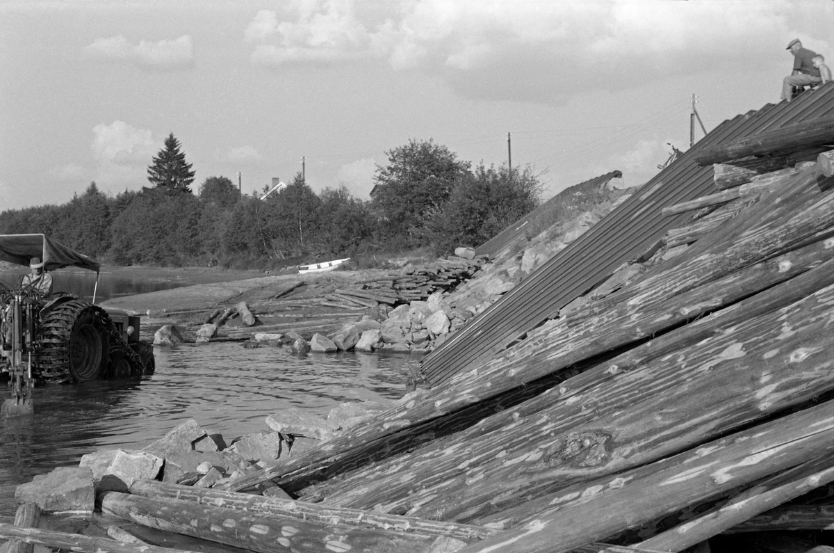 Mudring med beltegående traktorgraver ved utislagsplassen Tråstadsund i Glomma ved Kongsvinger i juli 1959. Vi ser deler av traktorgraveren i aktivitet på lav sommervannstand. Til høyre i bildet ser vi sjølve utislagsrampa - et skråplan lagd av parallelle jernbaneskinner der tømmeret fra lastebilene - i perioder med større vannføring - kunne skli ned i elva. Det ser for øvrig ut til å ha vært to slike ramper ved Tråstadsundet.
