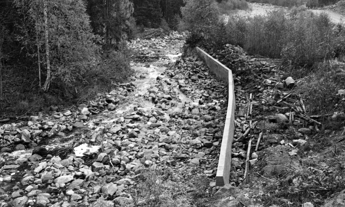 Nystøpt skådam i betong, bygget sommeren 1957, ved Kvernfossen i den delen av Gaukåa som ligger i Romedal allmenning, noen få hundrede meter ovenfor den nevnte åas utløp i innsjøen Rasasjøen. Fotografiet er tatt fra en bakkekam ned mot et steinete åløp, der det, i en slak yttersving, var reist en betongmur som skulle bidra til at fløtingsvirket holdt seg i vassdraget i stedet for å ende i omkringliggende krattskog, hvorfra det ville bli arbeidskrevende å bære det tilbake igjen. På landsida av skådammen ser vi stubber av eldre tømmer, antakelig fra en eldre skådam, en såkalt bukkedam.