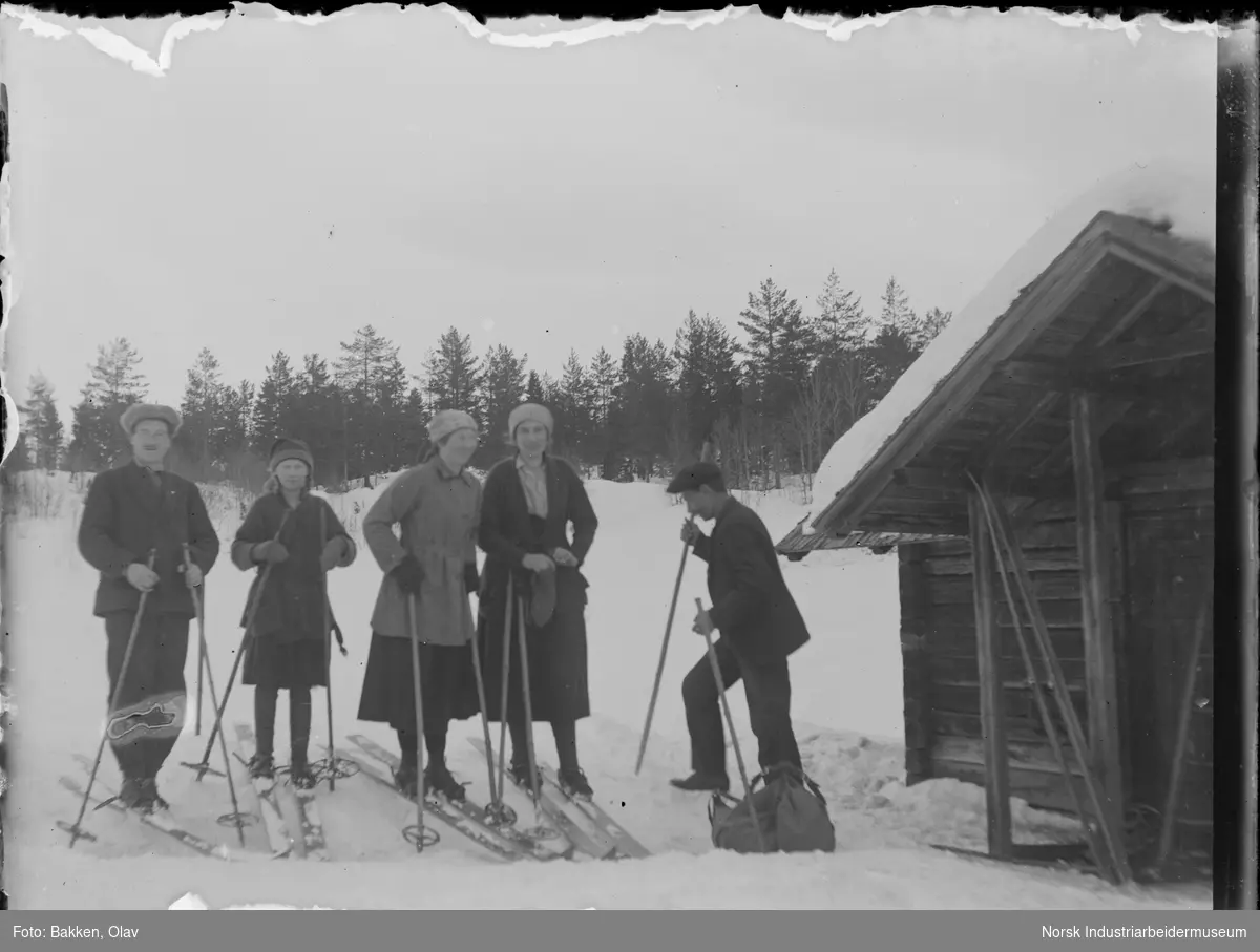 To menn, to kvinner og en jente stående ved hytte med ski på beina. Sekk liggende i snøen.