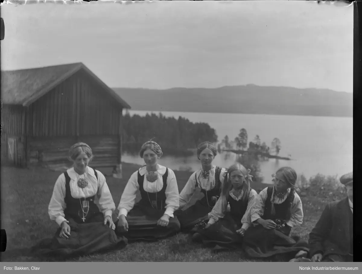 Tre kvinner og to jenter sittende i bunad på gresslette foran trebygg. Gutt delvis synlig til høyre. Utsikt mot Follsjø.