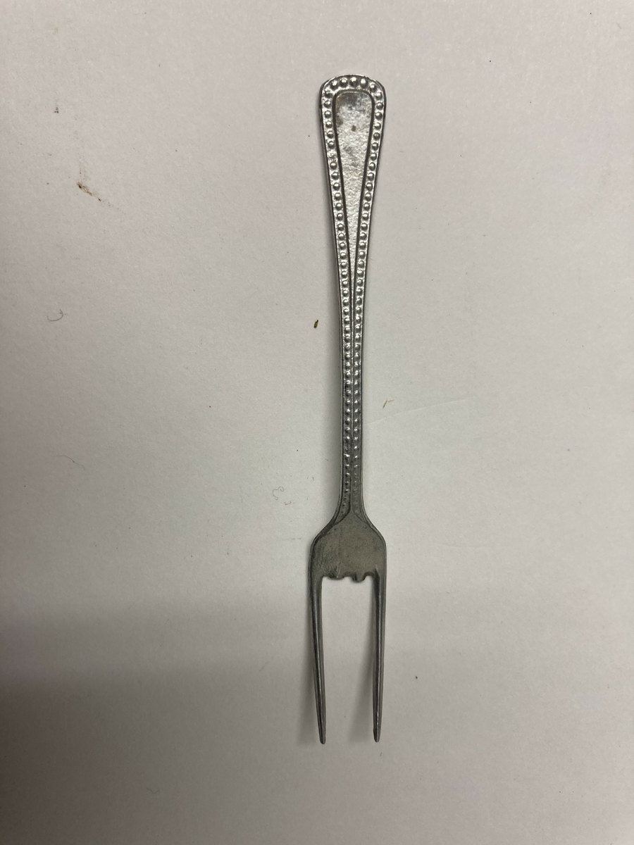 Gaffel til bestikk i ni deler til dokkestell. Gaffelen har en skade. Dokkestell ble gitt til jenter til lek som forberedelse til voksenlivet. 
