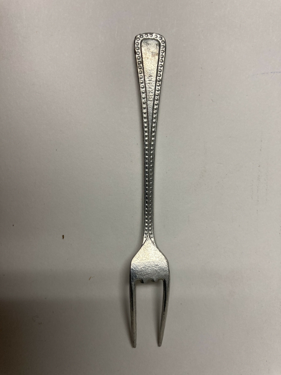 Gaffel til bestikk i ni deler til dokkestell. Gaffelen har en skade. Dokkestell ble gitt til jenter til lek som forberedelse til voksenlivet. 