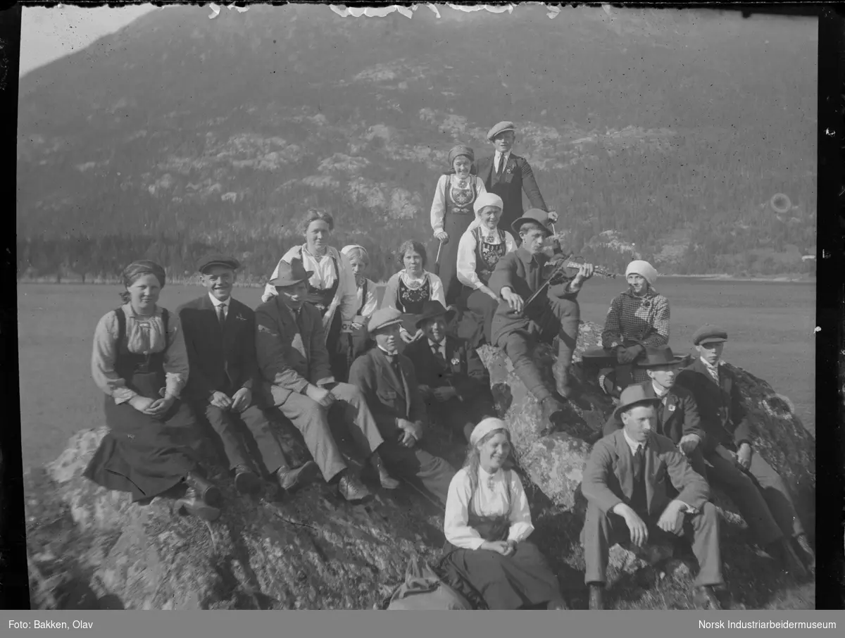 Gruppe mennesker på landtur. Alle ikledd dress og kjole/drakt sittende på en fjellknaus ved innsjø. Hølje Bakken spiller på fele.