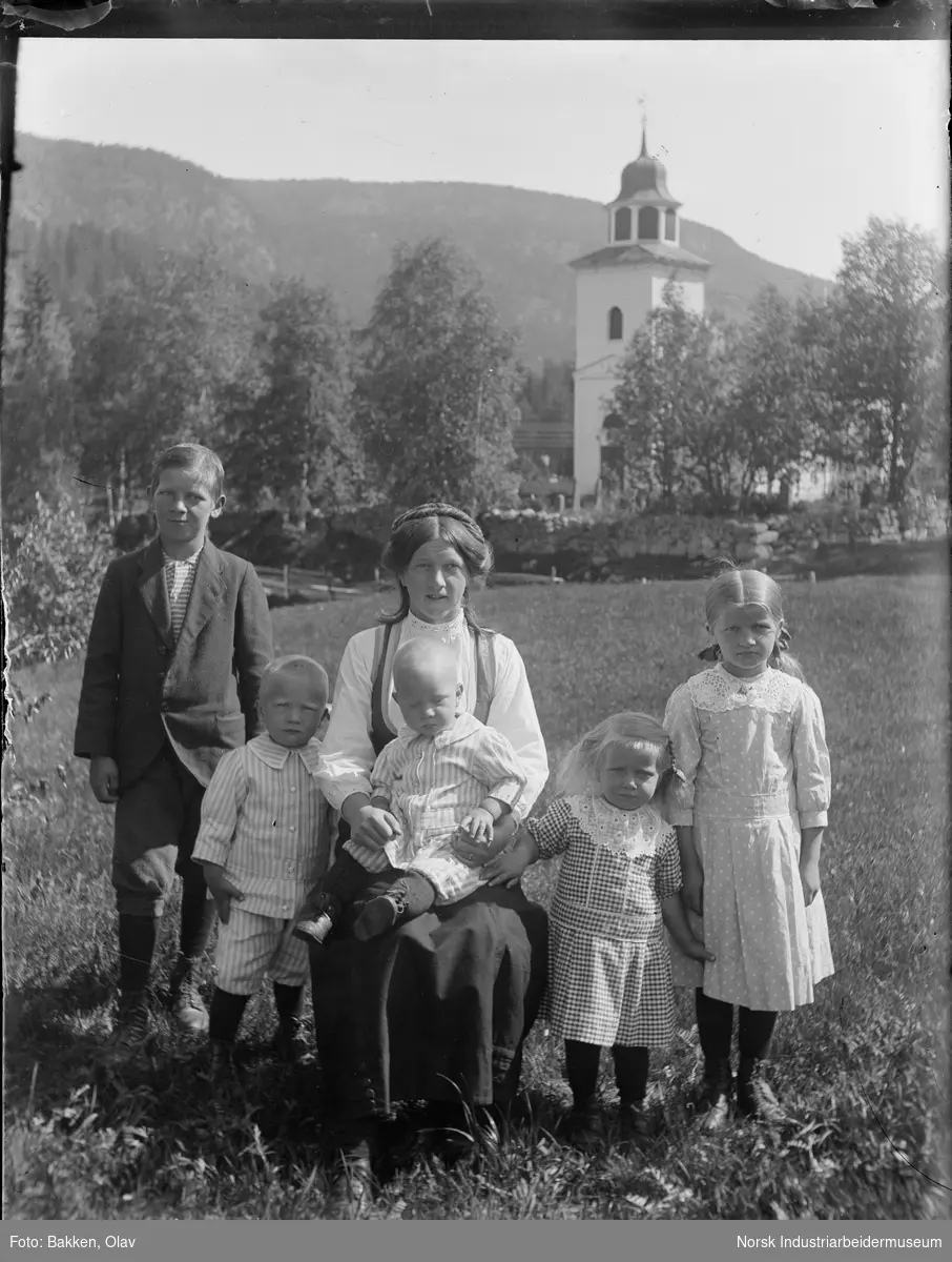 Kvinne med fem barn foran Gransherad kirke sittende på Prestegardsjordet. Kvinne sittende på stol, minste barnet sitter på fanget. Familien er ikledd pentøy.