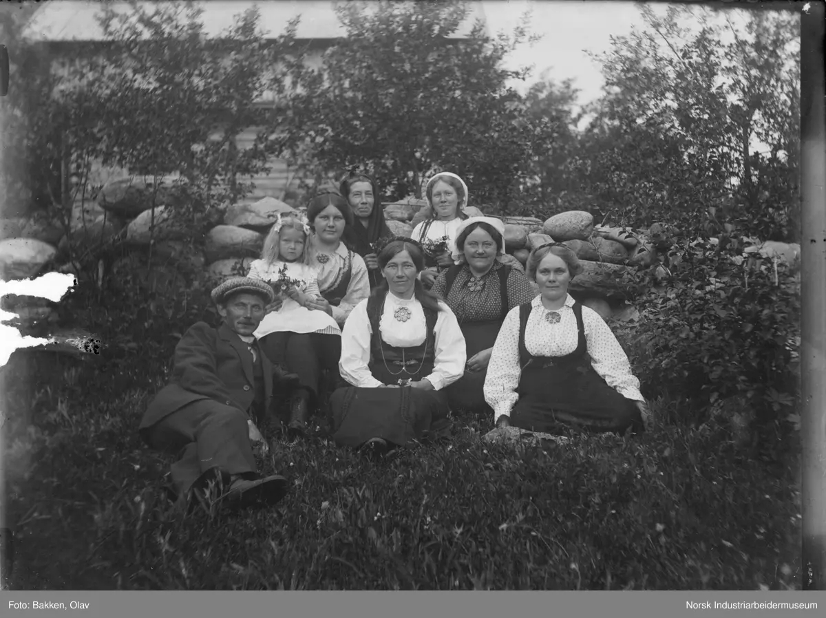 Gruppe mennesker sittende på gresset foran steinmur. Laftet bygg i bakgrunnen.