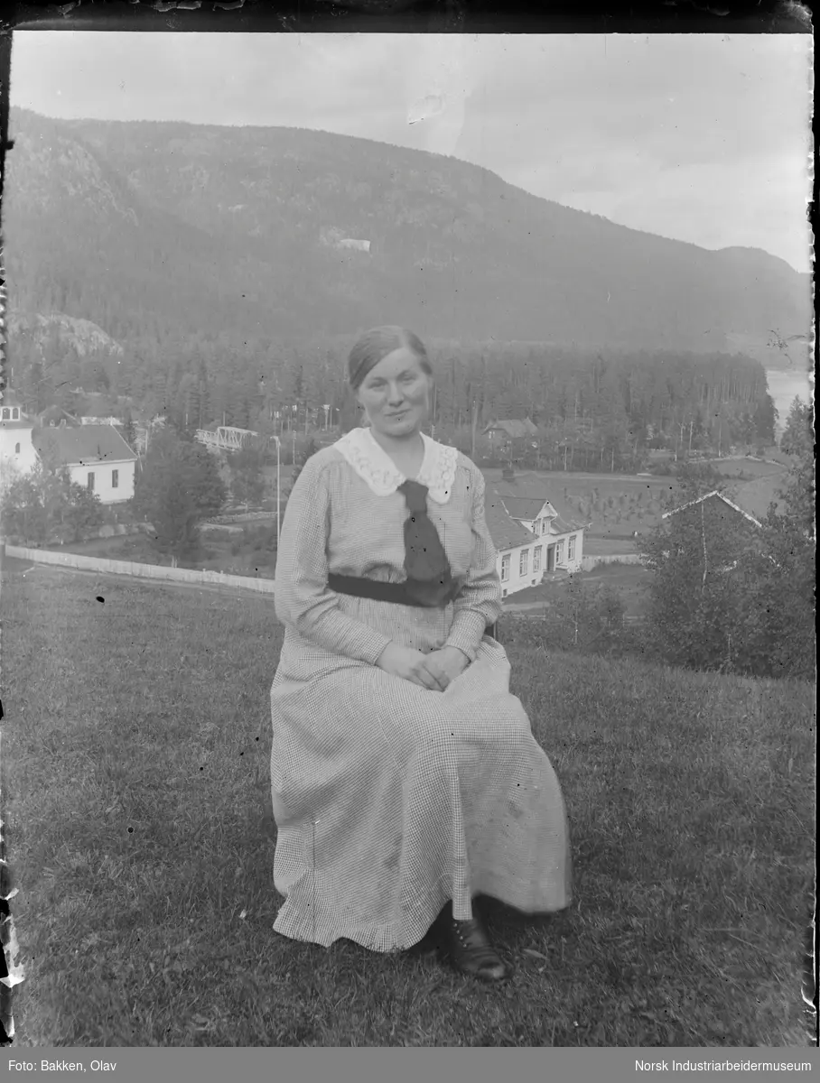 Portrett. Kvinne sittende utendørs. Tjenestepike i prestegården Abela Johannesen. I bakgrunnen Gransherad kirke og prestegården.