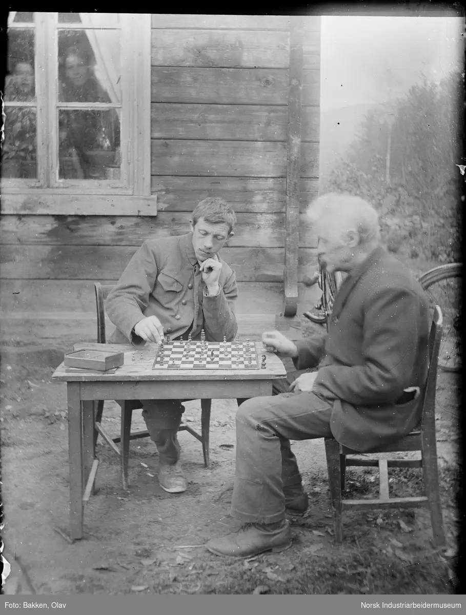 To menn sittende rundt bord utendørs med sjakkspill. To mennesker ser ut gjennom vinduet fra huset i bakgrunnen.