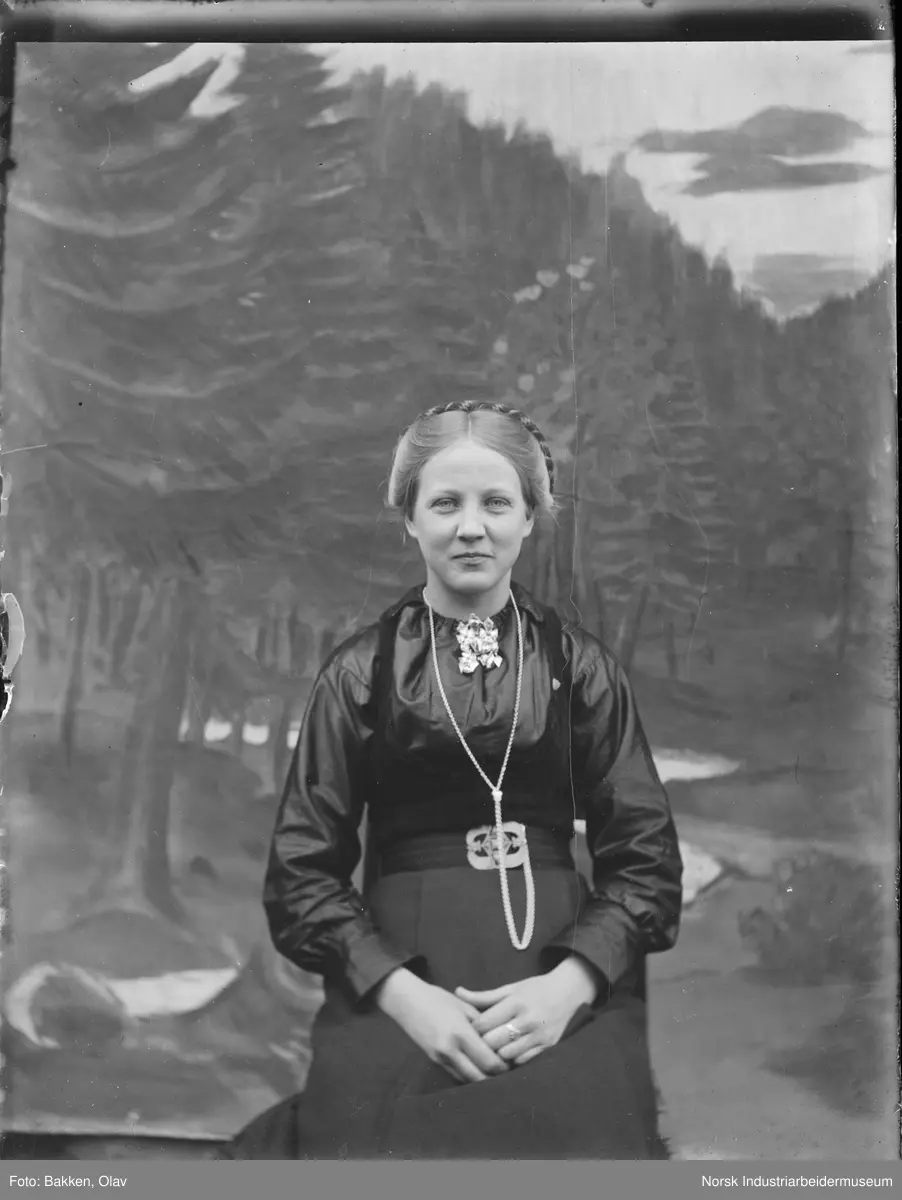 Portrett. Kvinne sittende foran malt bakgrunn.