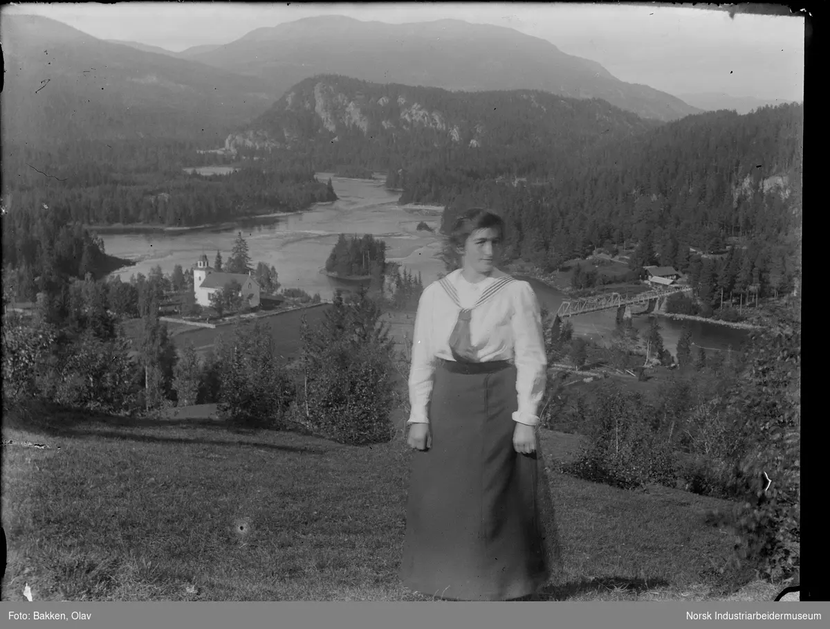 Portrett. Kvinne stående på gressbakke. Gransherad kirke og Gransherad bro i bakgrunnen.