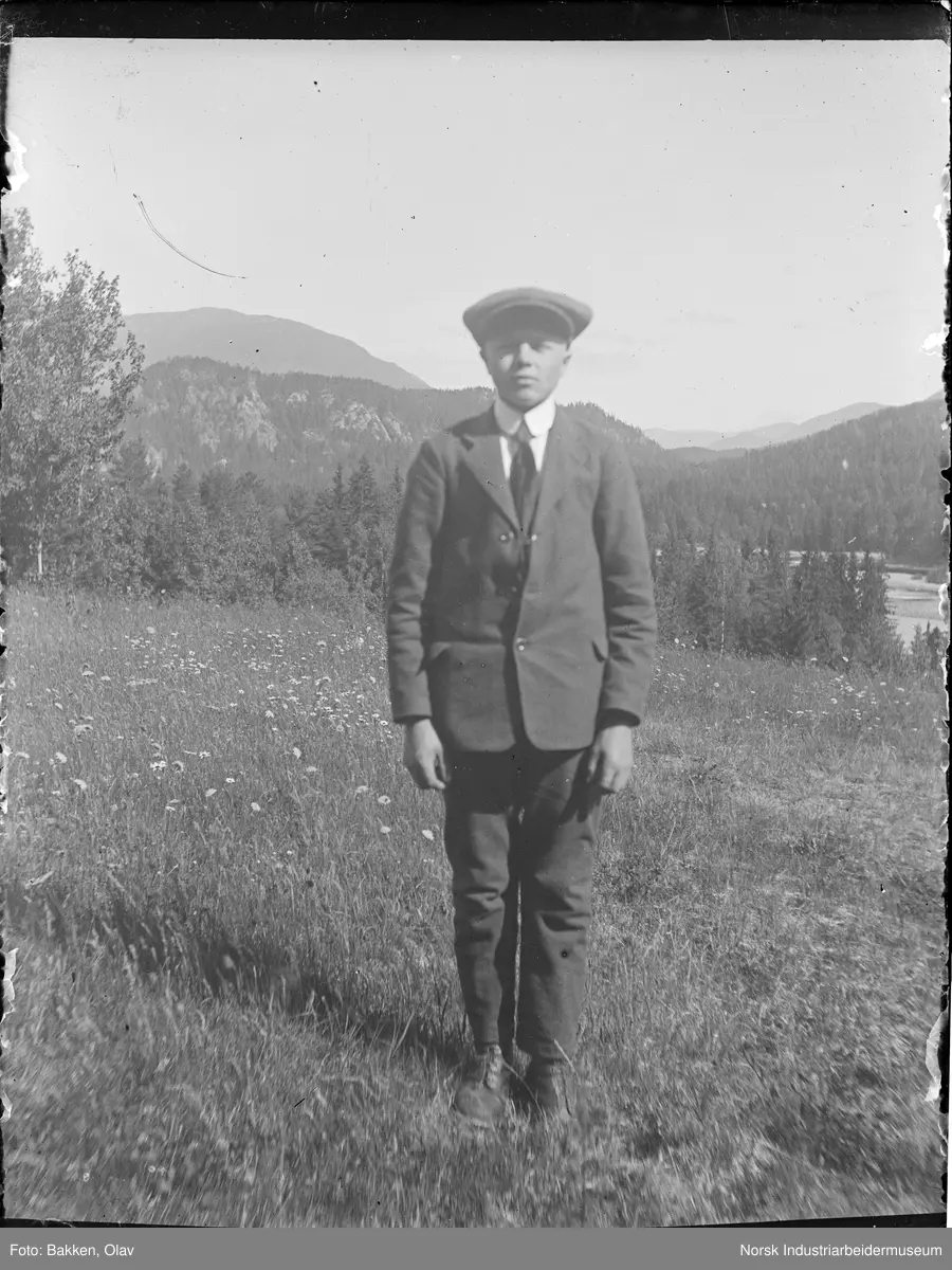 Portrett. Ung mann i dress ute på gressbakke. Fjell og vann i bakgrunnen.