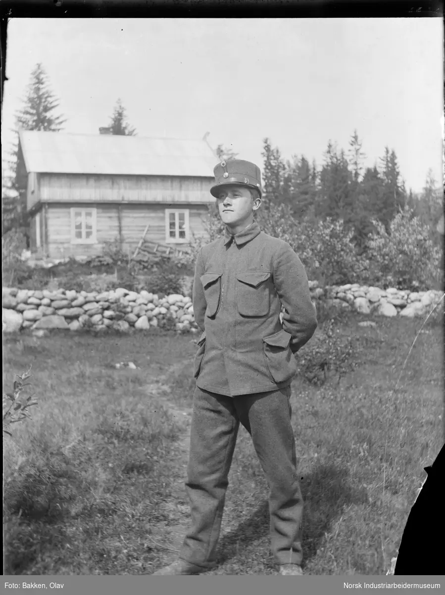 Portrett. Ung mann i soldat uniform på gressbakke. Bolig men utkraget andre etasje og steinmur i bakgrunnen.