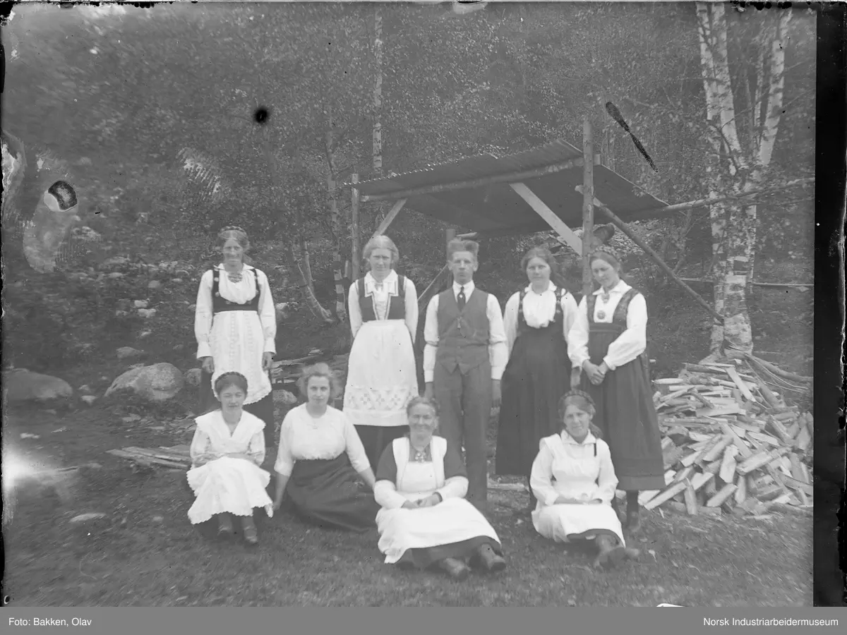 Gruppe mennesker oppstilt utendørs. Vedhaug og vedskjul i bakgrunnen.