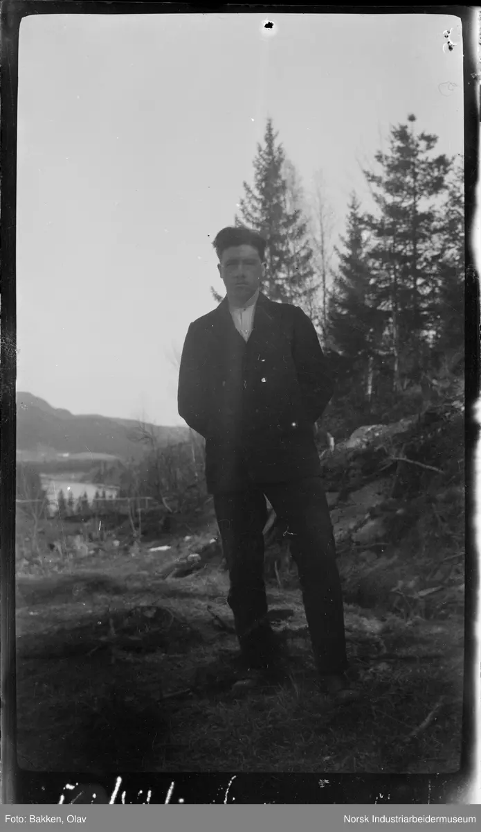 Portrett. Mann i skogsområde med vann i bakgrunnen. Kledd i dress.