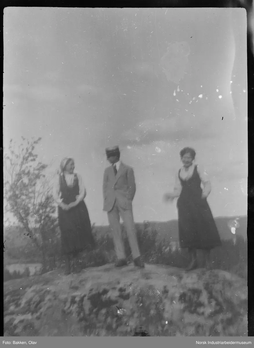 Mann og to kvinner stående på fjellknaus. Mannen i stråhatt og dress, kvinnene i drakt.