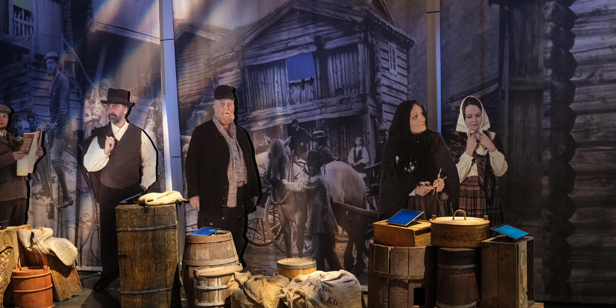 Utsnitt av utstillingen Tida og vegen. Bildet viser tønner, kasser og lignende sammen med figurer i 1800-tallskær foran veggbilde av et stabbur.