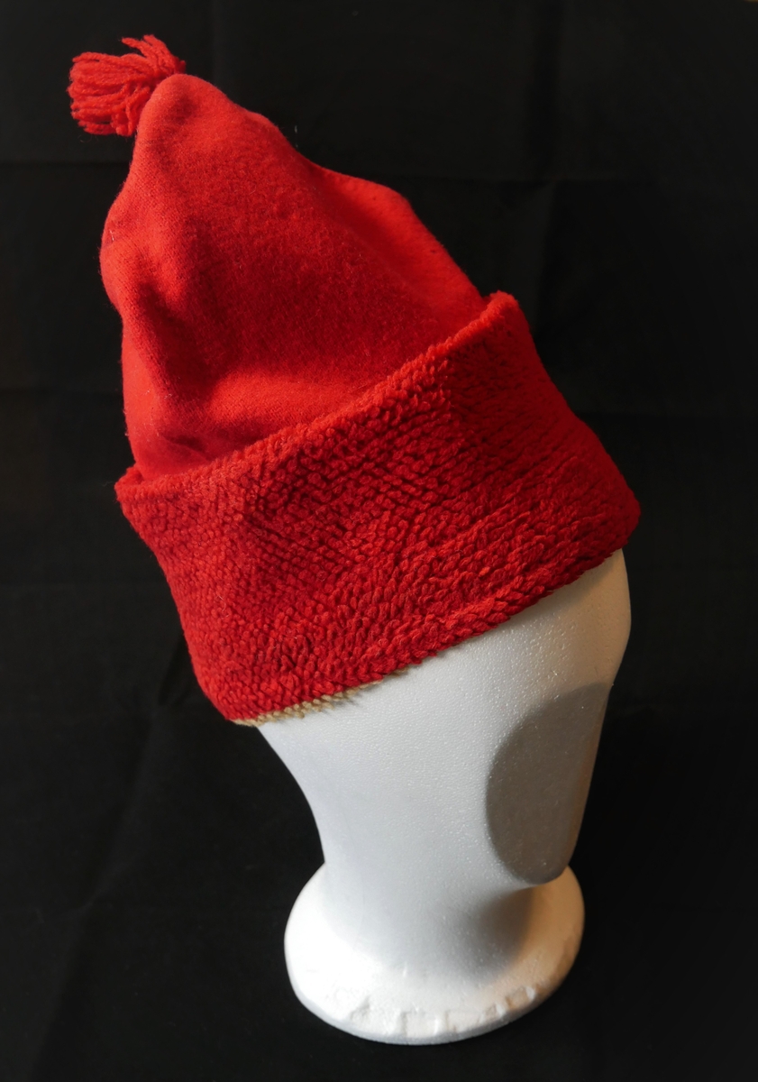 Ei strikka topphue, med dusk. To lag er sydd saman; det ytste er raudt glattstrikk, medan innsida er laga i flosstrikk (truleg) i kvitt og raudt. Brettekant nedst på hua, slik at den raude delen av innsida dannar flosskant på utsida. Laga er sydd saman i nedste kanten, samt i toppen der dusken er festa.