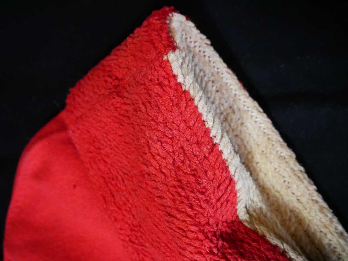 Ei strikka topphue, med dusk. To lag er sydd saman; det ytste er raudt glattstrikk, medan innsida er laga i flosstrikk (truleg) i kvitt og raudt. Brettekant nedst på hua, slik at den raude delen av innsida dannar flosskant på utsida. Laga er sydd saman i nedste kanten, samt i toppen der dusken er festa.