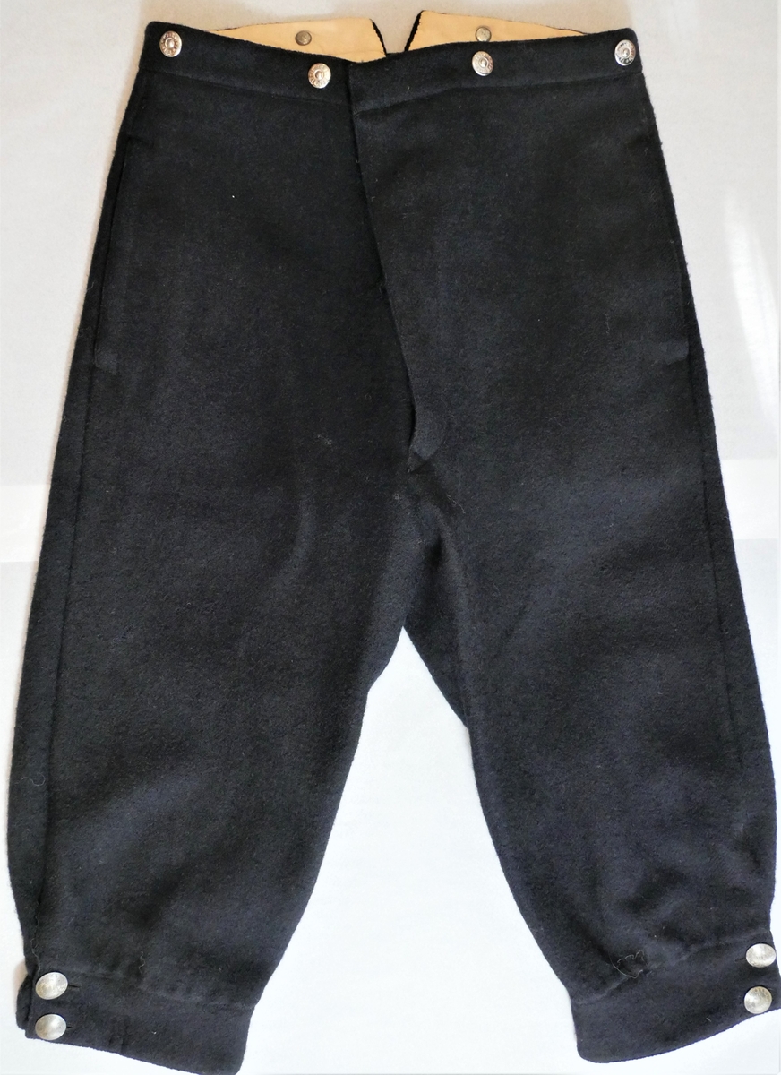 Ei kortbukse/knebukse til herre. Maskinsydd i vadmål, fôra i linninga og nedst i beina. Splitt med knepping nedst i beina - knappar av 50øringar (frå 1880, 1885, 1900 og 1901). Gylf med skjult knepping - tre treknappar, samt ei metallhempe øvst. Seks knappar (nyare) i linninga, til feste av bukseselar (to på baksida og to på kvar side av gylfen framme). Stikklommer i sidene. Splitt i linninga bak, med reimer og spenne til stramming.