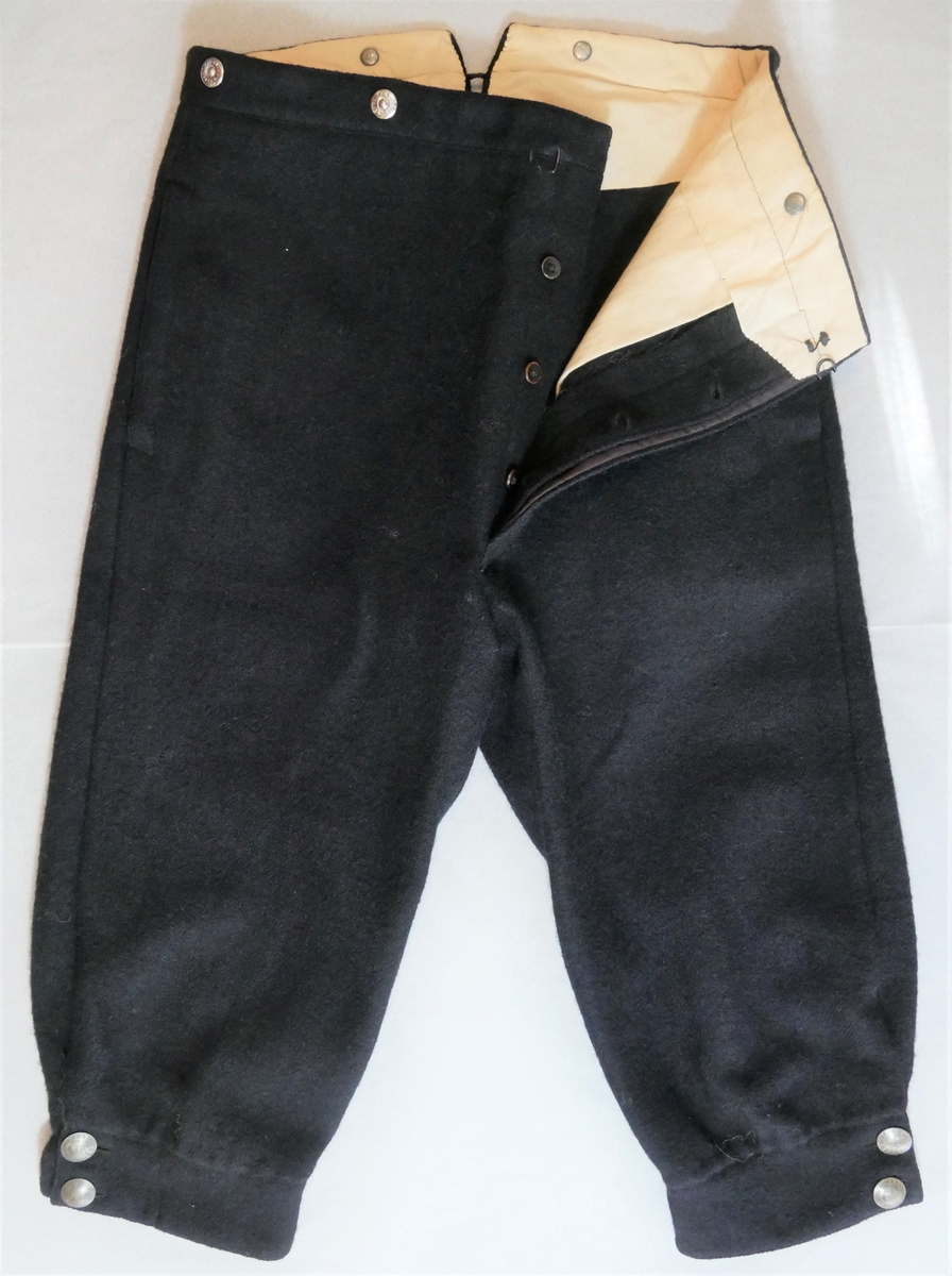 Ei kortbukse/knebukse til herre. Maskinsydd i vadmål, fôra i linninga og nedst i beina. Splitt med knepping nedst i beina - knappar av 50øringar (frå 1880, 1885, 1900 og 1901). Gylf med skjult knepping - tre treknappar, samt ei metallhempe øvst. Seks knappar (nyare) i linninga, til feste av bukseselar (to på baksida og to på kvar side av gylfen framme). Stikklommer i sidene. Splitt i linninga bak, med reimer og spenne til stramming.