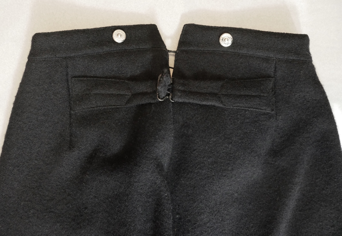 Ei kortbukse/knebukse til herre. Maskinsydd i vadmål, fôra i linninga og nedst i beina. Splitt med knepping nedst i beina - knappar av 50øringar (frå 1880, 1885, 1900 og 1901). Gylf med skjult knepping - tre treknappar, samt ei metallhempe øvst. Seks knappar (nyare) i linninga, til feste av bukseselar (to på baksida og to på kvar side av gylfen framme). Stikklommer i sidene. Splitt i linninga bak, med reimer og spenne til stramming.