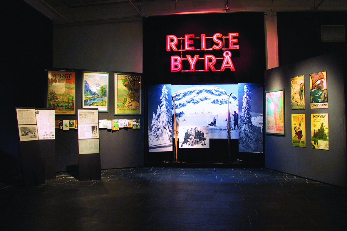 Fra utstillingen, plakater, vilder og kart stilt ut på vegg. Ski stilt ut med vinterlandskap i bakgrunn. På vegg står det "reisebyrå" i neonlys.