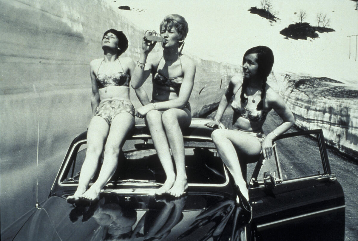 Fotografi av tre unge demer i bikini som sitter på taket av en bil. En drikker fra en glassflaske. Rundt dem er det snø.