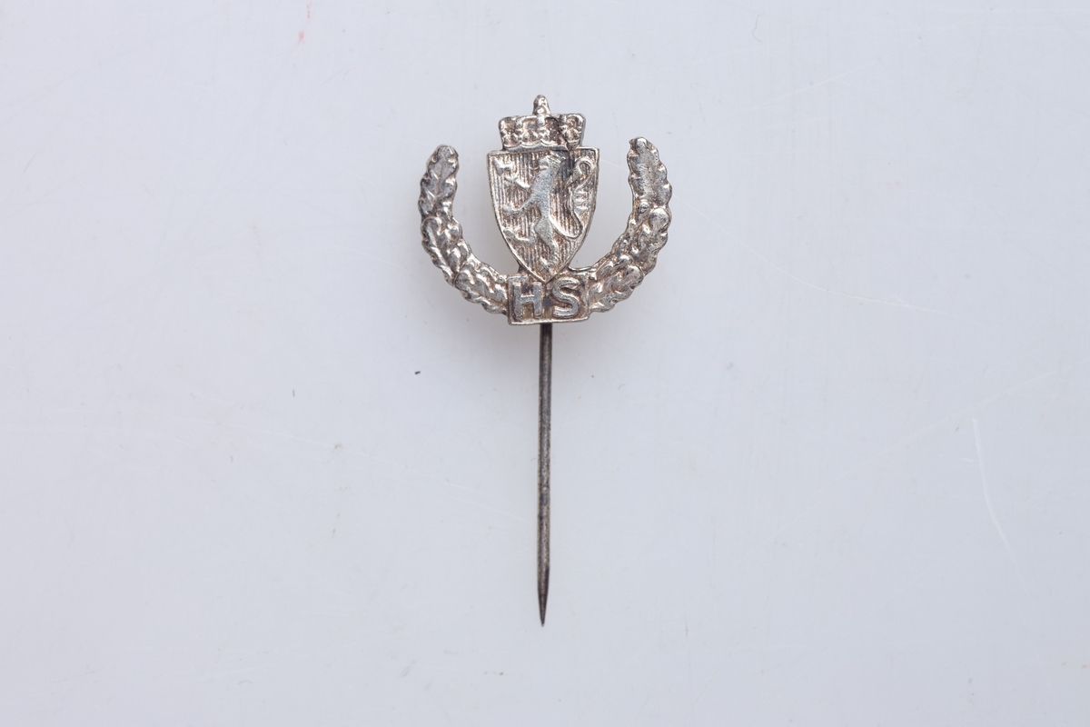 Nål for deltakelse i de norske hjemmestyrkene. Nålen viser Riksvåpnet over H S, og ekeløv oppover langs sidene. Støpt merke med påloddet nål.
