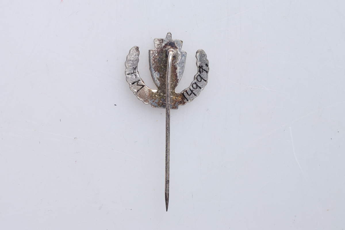 Nål for deltakelse i de norske hjemmestyrkene. Nålen viser Riksvåpnet over H S, og ekeløv oppover langs sidene. Støpt merke med påloddet nål.