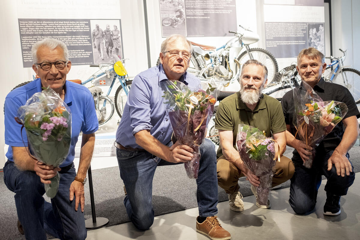 Fire menn sitter på kne med hver sin blomsterbukett foran utstillingen Basse i 100.
