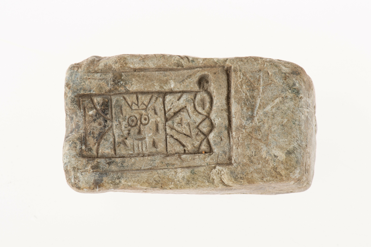 På ena bredsidan streckfigurer (bl.a. ett ansikte med en krona), på den motsatta sidan en hemisfärisk urgröpning. Även en oval urgröpning på ena smalsidan. I huvudliggaren upptagen som gjutform för rektangulärt spänne.