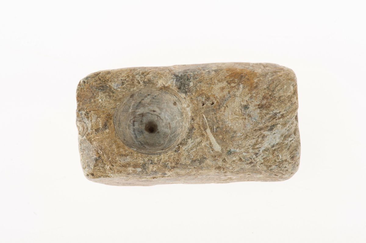 På ena bredsidan streckfigurer (bl.a. ett ansikte med en krona), på den motsatta sidan en hemisfärisk urgröpning. Även en oval urgröpning på ena smalsidan. I huvudliggaren upptagen som gjutform för rektangulärt spänne.