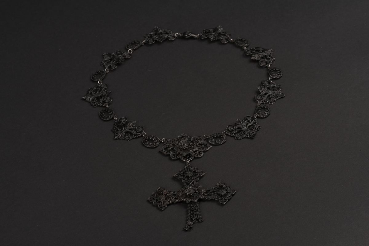 Svart halsband av gjutjärn.
Halsbandet består av omväxlande större och mindre utsirade länkar i nygotisk stil, som är sammanfästa med runda små länkar. Mitt fram hänger ett större likformat kors, som är något skadat. Spännet är utformat som två händer.