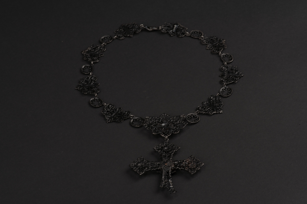 Svart halsband av gjutjärn.
Halsbandet består av omväxlande större och mindre utsirade länkar i nygotisk stil, som är sammanfästa med runda små länkar. Mitt fram hänger ett större likformat kors, som är något skadat. Spännet är utformat som två händer.