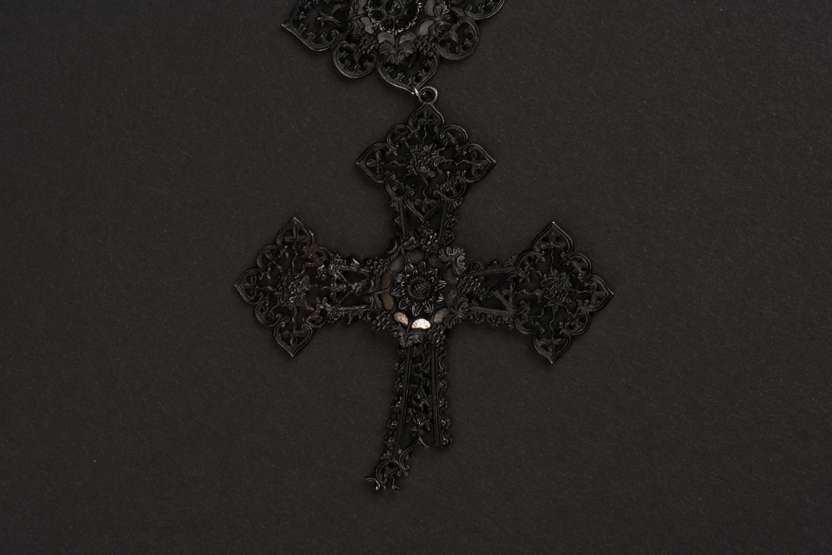 Svart halsband av gjutjärn.
Halsbandet består av omväxlande större och mindre utsirade länkar i nygotisk stil, som är sammanfästa med runda små länkar. Mitt fram hänger ett större likformat kors, som är något skadat. Spännet är utformat som två händer.